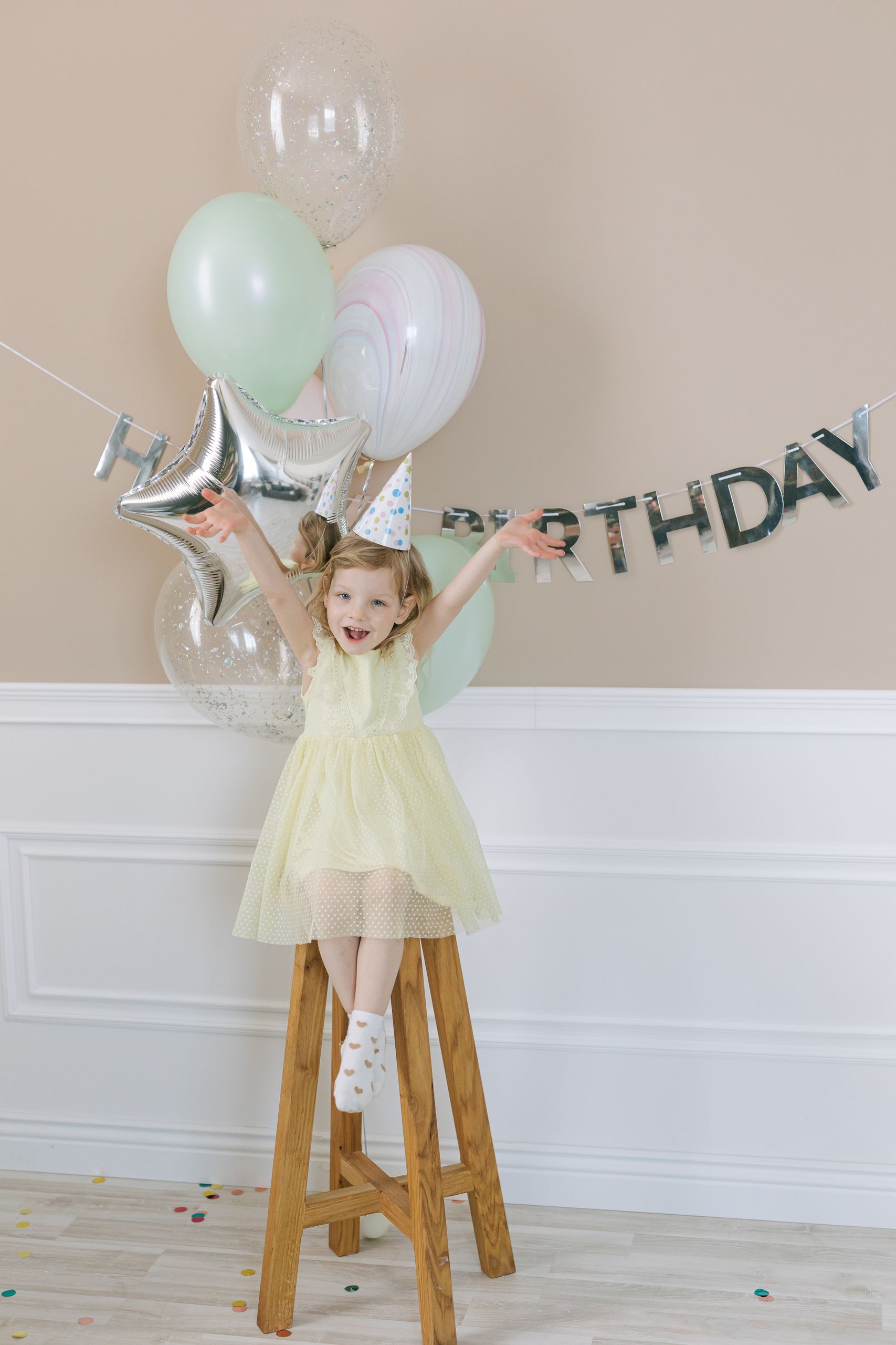 A little girl joyfully celebrates her birthday indoors with balloons and a party hat, sitting on a stool.
