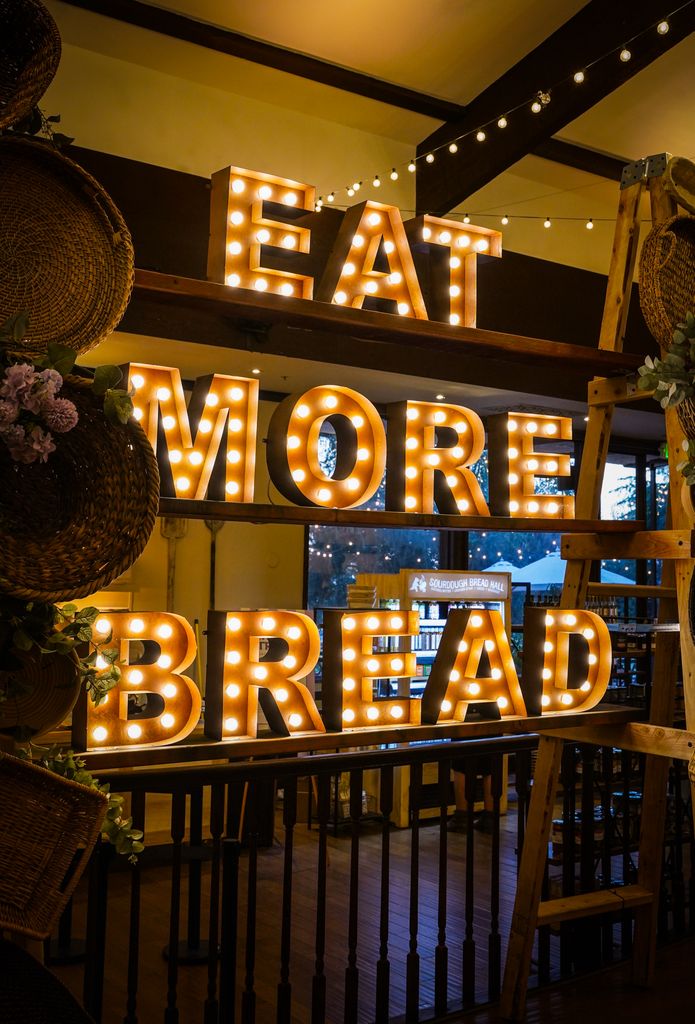 Brightly lit sign reading 'Eat More Bread' in a cozy indoor setting with decorative baskets.